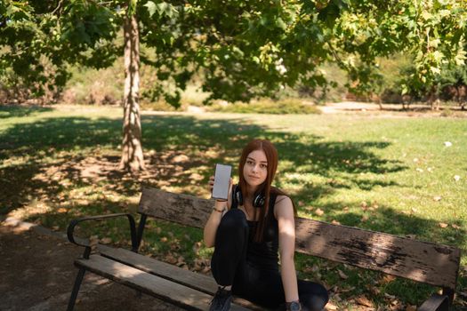 Young athletic female in black activewear sitting on park bench in summer and showing empty screen of smartphone