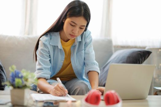 Young asian woman surfing the internet using laptop and holding book while sitting on couch at home. Young asian women studying online at home. Online connection and online learning concept...