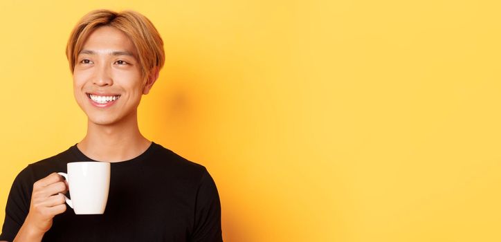 Close-up of happy handsome young asian guy with blond hair, looking dreamy and smiling while drinking coffee or tea, standing over yellow background.