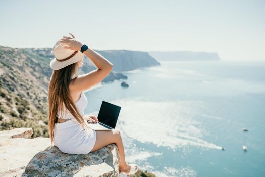 Successful business woman in yellow hat working on laptop by the sea. Pretty lady typing on computer at summer day outdoors. Freelance, travel and holidays concept.