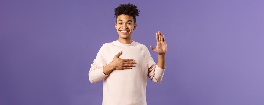 Portrait of young happy, joyful hispanic man making honest statement, promise to tell only truth, hold one hand on heart and another raised, smiling sincere, standing purple background.