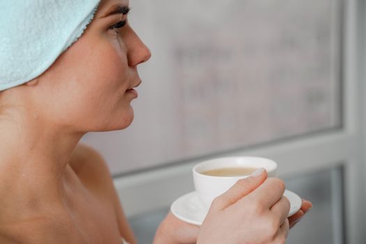 Young serene relaxed woman in spa bath towel drinking hot beverage tea coffee after taking shower bath at home. Beauty treatment, hydration concept