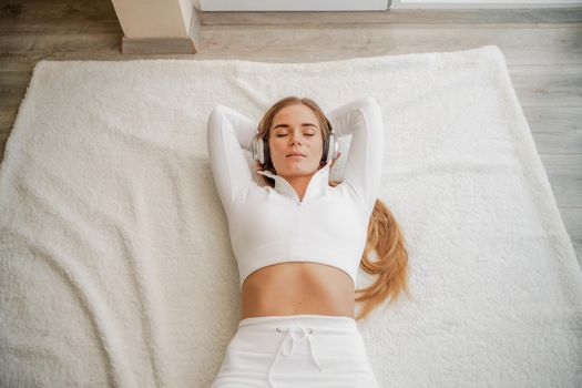 Top view portrait of relaxed woman listening to music with headphones lying on carpet at home. She is dressed in a White tracksuit