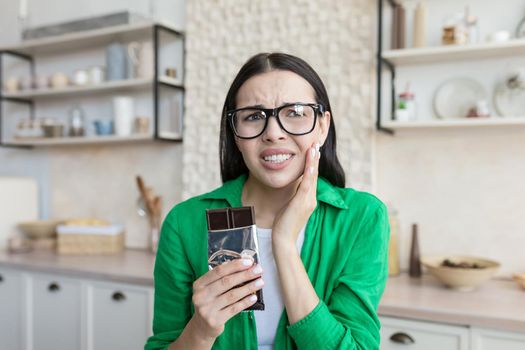 Toothache. Portrait of a young beautiful woman at home in glasses and a green shirt who has a toothache from sweets. He holds a bar of chocolate in his hands, holds his cheek, feels severe pain.