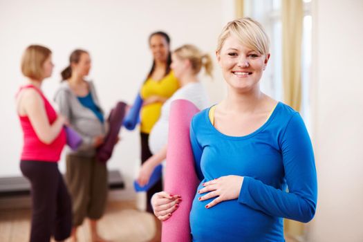 Pilates for pregnancy. A young blonde pregnant woman in a gym holding an exercise mat with a group of women in the background
