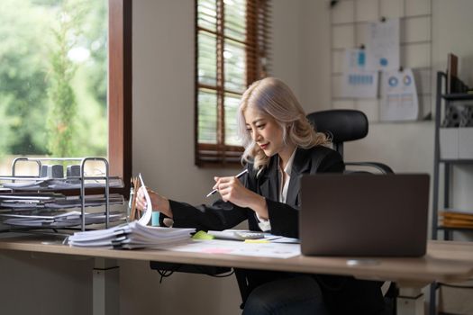 Young asian businesswoman working with laptop, financial charts and graphs reviewing data in paper documents making money work at office desk.