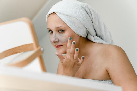 Young woman wrapped in towel puts gray cosmetic clay on her face while looking in mirror. Close-up