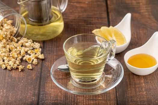 Relaxing chamomile tea in glass cup with lemon slices and honey next to it on wooden table.