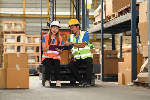 Warehouse workers, sitting on forklift truck and checking newly arrived goods in large warehouse.