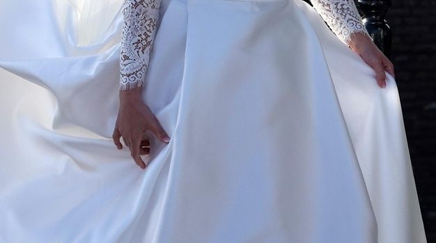 The hands of a bride. She lifts her long skirt to walk. The sleeves of her white dress are made of lace.