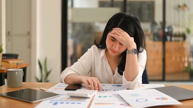 Tired asian woman employee sitting at her office desk, holding her head in hands. Overworking, stress, depression concept.