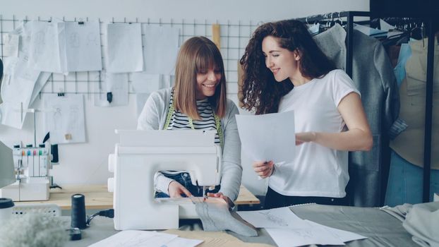 Professional seamstress is stitching fabric with sewing machine then adjustig equipment while her female colleague is showing her sketches. Light studio with tailoring items in background.