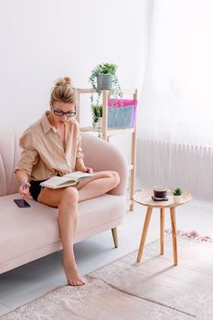 A serious modern slender woman of 30-40 years old, in casual clothes and glasses, in white wired headphones, sits on a comfortable sofa in a bright interior, writes in a diary. Vertical. Copy space