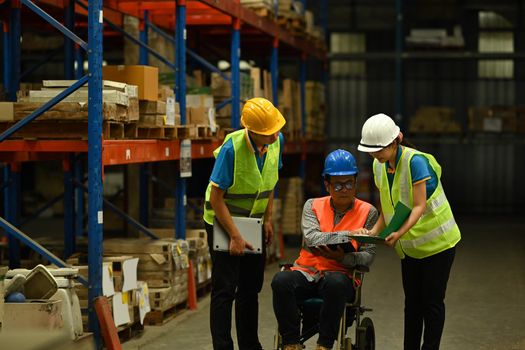 Male manager in wheelchair and young workers inspecting stock tick and cardboard stock product in a large warehouse.
