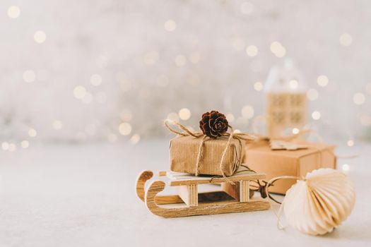 Eco Natural Christmas gift on toy wooden sled on white background with glowing light bokeh, copy space. New year card. Xmas decoration with house, Christmas tree and snow