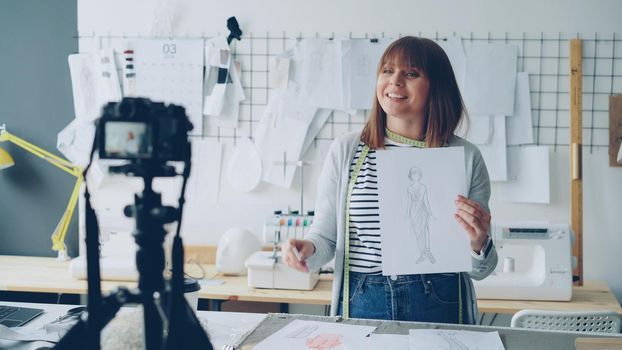 Young blogger seamstress is recording video for instagram about designing women's clothing. She is greeting followers, showing sketch, speaking and gesturing. Professional blogging concept.