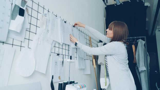 Young creative clothes designer is looking at sketches then hanging them on wall above studio table beside other drawings. Loft style studio and fashionable garments are visible.