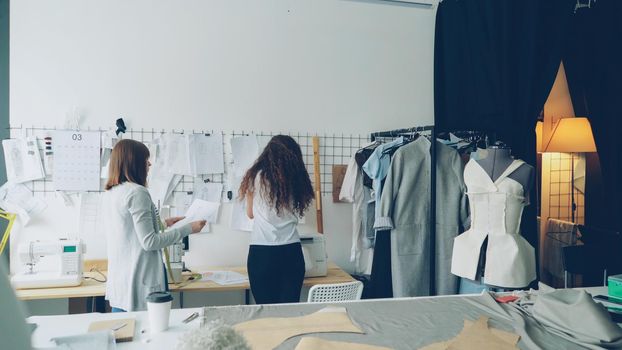 Young female entrepreneurs clothing designers standing in front of wall with hanging sketches, discussing drawings and placing images with clips. Creative team working together concept.