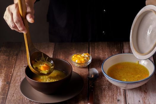 woman serving chicken soup with a wooden spoon in a brown bowl on a wooden table