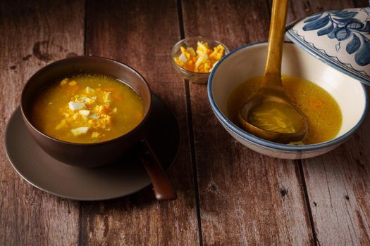 chicken noodle soup in a white soup tureen and a brown bowl with a plate and spoon on a wooden table