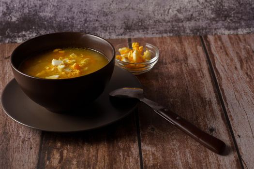 chicken noodle soup in a brown bowl with spoon and hard-boiled egg on a wooden table