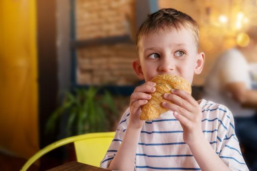 Portrait of a little boy, blond of European appearance in cafe on summer day. The child eats a croissant for breakfast. The concept of baking unhealthy food. Baking, bakery, malnutrition for children