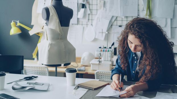 Young female artist fashion designer is drawing women's garment sketch with pencil at table in modern studio. Mannequin, sewing items, take-away coffee are visible.