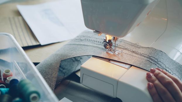 Close-up of modern electric sewing machine working stitching piece of textile. Girl's manicured hand, sketches on paper and colorful sewing threads are visible.