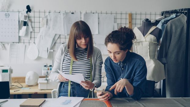 Young clothing design entrepreneurs are looking at sketches and checking smartphone while working at sewing desk. Attractive women are discussing fashion industry in friendly manner.