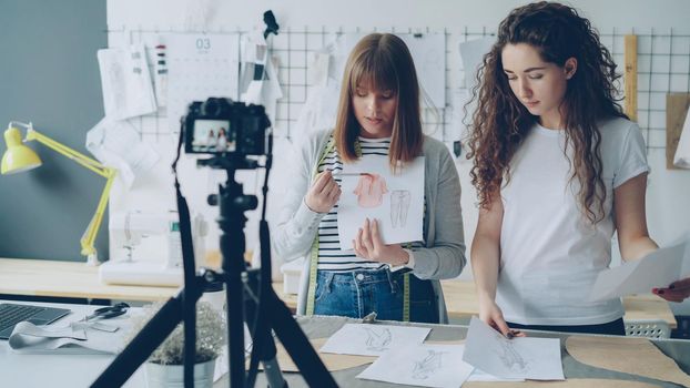 Young attractive women fashion bloggers recording video blog about ladies'clothes on camera and talking to followers in modern studio. Many garment sketches are visible.