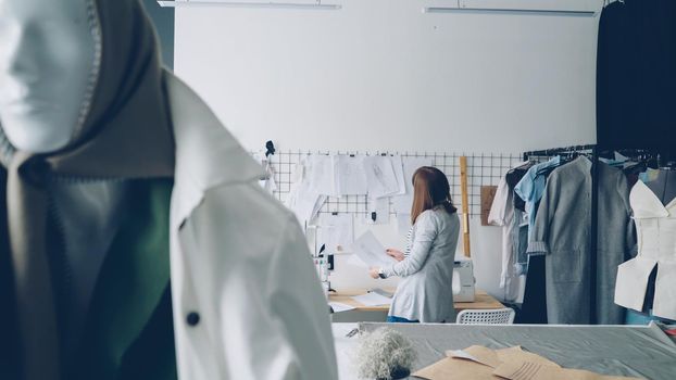 Attractive young woman clothing designer is looking at garment sketches hanging on wall then choosing new drawings. Mannequin dresses in trendy clothes is in foreground.