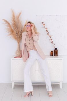 a stylish woman in glasses, of 30-40 years old with blond hair stands in a light pink jacket and white trousers in the interior of a white room, looks to the side. Copy space. Vertical