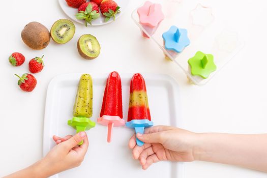 Two childrens hands, take multi-colored homemade kiwi and strawberry ice cream from a white square plate, on a white table, top view