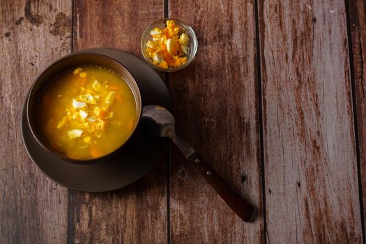 chicken noodle soup in a brown bowl with spoon and hard-boiled egg on a wooden table