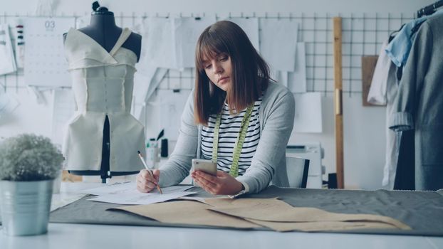 Creative clothing designer is looking at smartphone screen and drawing sketch while working in modern tailor shop at sewing table. Woman is busy and involved in process.