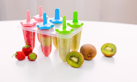 A plastic mold with multi-colored homemade kiwi and strawberry ice cream stands on a white table by the window in daylight. Near kiwi and strawberry