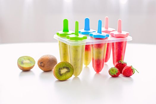 A plastic mold with multi-colored homemade kiwi and strawberry ice cream stands on a white table by the window in daylight. Near kiwi and strawberry