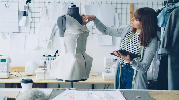 Young female fashion designing entrepreneur is working with tablet while checking clothing pinned to tailoring dummy. Modern technologies in clothes manufacturing concept.
