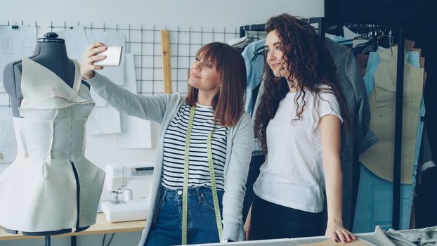 Two cheerful female clothing designers are making funny selfie with smart phone while standing beside clothed mannequin in studio. Attractive women are smiling and posing.