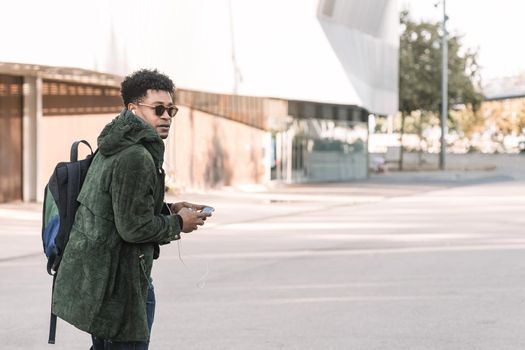 african american young male with earphones walking by the city and listening music with a mobile phone, technology and lifestyle concept using internet electronic device outdoors