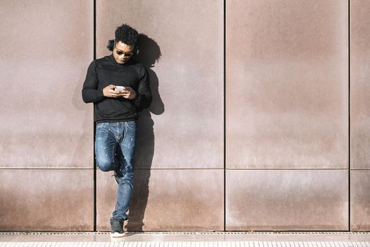 cool african black man with casual modern clothes and headphones listening music of his phone leaning against wall, technology and lifestyle concept