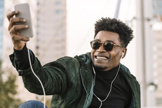 handsome young african black man with sunglasses making a video call with smartphone and earphones while sitting in the city, lifestyle and technology concept using internet electronic device