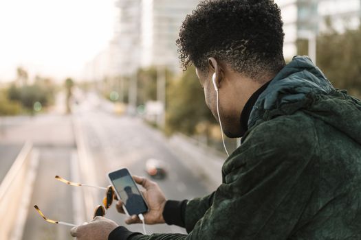 view from behind of a young african black man with sunglasses making a video call with smartphone and earphones, city lifestyle and technology concept using internet electronic device