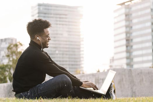 smiling young black student laughing with earphones making a video call with the computer sitting on the lawn in the city at sunset, lifestyle and technology concept using internet electronic device