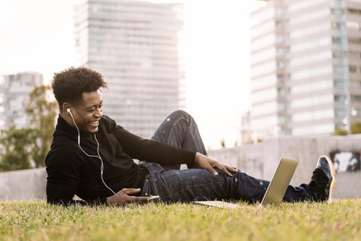 smiling young black man laughing with white earphones looking to the laptop lying on the lawn in the city at sunset, lifestyle and technology concept using internet electronic device