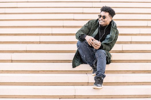 young black man with sunglasses in casual modern clothes sitting on stairs, listen to music of his phone in earphones while smile, technology and lifestyle concept