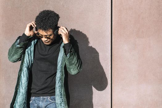cool african black man with casual modern clothes and headphones listening music leaning against wall, technology and lifestyle concept