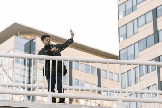 happy black man posing for selfie on smartphone camera while listen music in headphones or making video call outdoors in the city, technology and lifestyle concept using internet electronic device