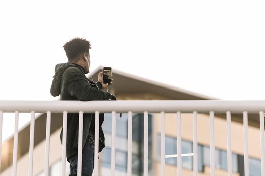 african black man in earphones taking a photo at sunny street of the city with his phone, selfie for the social networks, technology and lifestyle concept using internet electronic device
