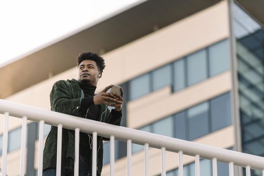 portrait of a young cool african black man listening to music with earphones and mobile phone while leaning on a handrail outdoors in the city, technology and lifestyle concept